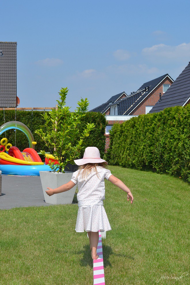 Ein Mädchen balanciert auf ihrem Balancierbalken im Garten und trägt dabei ein weißes Kleid. Im Hintergrund sieht man den blauen Himmel und einen Kinderpool. 