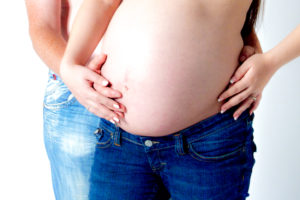 Eine schwangere Frau zeigt ihren nackten babybauch., denn sie trögt nur eine Jeans. Ihr mann, ebenfalls in jeans steht hinter ihr und hält den babybauch von hinten mit der einen und sie mit dem anderen in der hand.