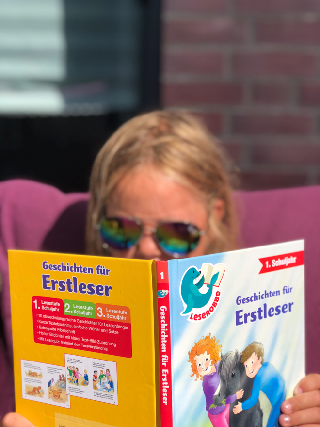 Kind liest mit einer Sonnenbrille ein Erstlesebuch. Lesen macht Spaß und das sollten wir unseren Kindern behalten. Mit dem Sommer-Lesepass macht es noch viel mehr Freude. Nicht nur in den Sommerferien.