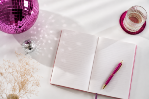 Aufgeschlagenes Notizbuch mit pinkfarbenem Stift und Discokugeln auf weißem Hintergrund